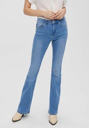 Kobieta ubrana w jasnoniebieskie jeansy z rozszerzanymi nogawkami, biały top i beżowe botki z noskiem w szpic, stojąca z rękami złożonymi na wysokości talii.