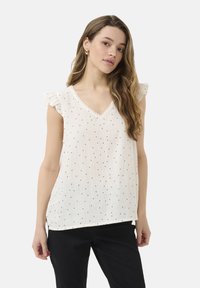 Jeune femme aux longs cheveux ondulés portant un haut blanc sans manches à col en V avec de petits points noirs et un pantalon noir, debout devant un fond blanc.