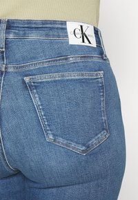 Jeans de mezclilla azul de tiro alto con un bolsillo trasero visible, que presentan una etiqueta blanca con el logotipo de CK en la cintura y un ligero desgaste.