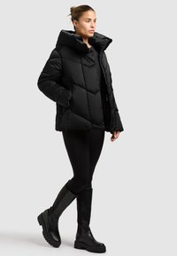 Schwarze Steppdaunenjacke mit hohem Kragen, Rautensteppung und Vorderentaschen, kombiniert mit schwarzen Leggings und kniehohen schwarzen Stiefeln.