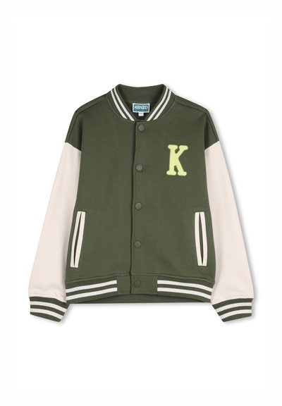 Veste universitaire verte avec manches crème, poignets et ourlet côtelés, arborant un écusson jaune en forme de "K" sur la poitrine et une fermeture à boutons pression.