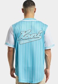 Tricou cu dungi albastre deschise și mâneci albe. Prezintă logo-ul "Kani" pe spate cu "est. 1989." Fabricat dintr-un material respirabil cu o ajustare lejeră.