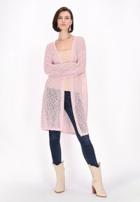 Helles rosa offen gestrickte Strickjacke mit langen Ärmeln, kombiniert mit einem beigen Oberteil, dunklen Denim-Jeans und cremefarbenen Cowboy-Stiefeln.
