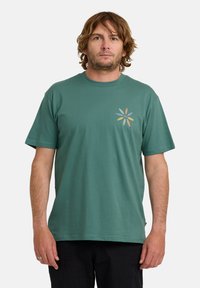 Teal bomulds t-shirt med kort ærme design og et lille flerfarvet blomstermotiv på venstre bryst.