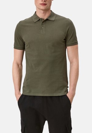 Uomo con polo verde oliva a maniche corte e pantaloni neri, con le mani nelle tasche, su uno sfondo chiaro e uniforme.