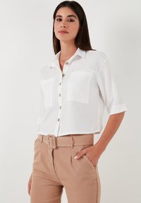 White, short-sleeve, button-up shirt with two chest pockets and a textured fabric, paired with light brown belted pants.