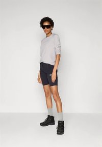 Pessoa usando óculos de sol oversized, camisa de manga longa cinza clara, calções pretos, meias cinzas e botas de caminhada pretas, de pé junto a uma parede simples.