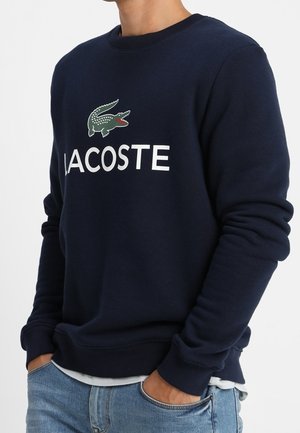 Osoba ubrana w granatową bluzę z zielonym logo krokodyla i białym napisem "LACOSTE", w połączeniu z jasnoniebieskimi dżinsami, ręce w kieszeniach.