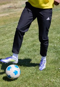 Svarta träningsbyxor med vita strumpor och blå och silverfärgade fotbollsskor, sparkar en fotboll på grönt gräs.