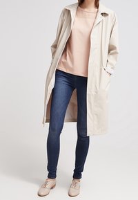 Beige voděodolný trench coat se jednoduchým designem, spárovaný se světle růžovým topem, modrými skinny džínami a béžovými botami.