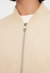 Μπεζ μπουφάν μπόμπερ με ριμπ κολάρο, ασημένιο φερμουάρ και απαλή υφή. Κοντινή λήψη αναδεικνύει το υλικό και το σχέδιο.