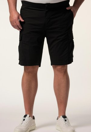 Mand iført sorte cargoshorts og hvide sneakers med sorte detaljer, stående med hænder nær lommerne mod en ensfarvet baggrund.