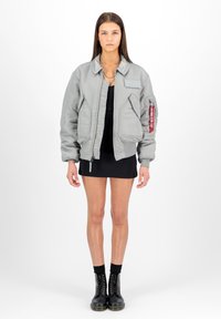 Giacca bomber grigia con una texture lucida, caratterizzata da una zip frontale, tasche laterali e un'etichetta rossa sul braccio sinistro, indossata sopra un abito nero.