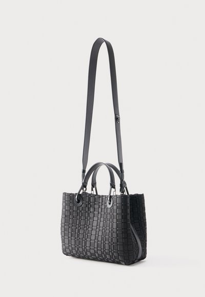 Μαύρη υφαντή τσάντα με δύο κοντά χερούλια και ένα μακρύ ρυθμιζόμενο ιμάντα ώμου, που στέκεται όρθια σε απλό φόντο.