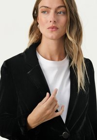 Czarna aksamitna marynarka z klapami, zapinana na jeden guzika, z długimi rękawami, noszona na białym t-shircie, charakteryzująca się gładką fakturą.