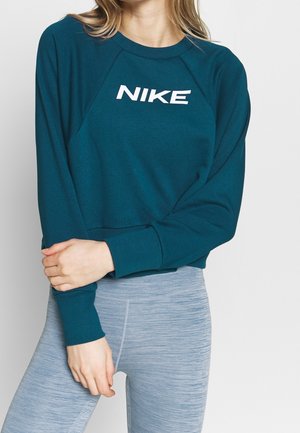 Kobieta w turkusowej bluzie Nike i jasnoszarych legginsach sportowych, stojąca z założonymi rękami na tle białego tła.