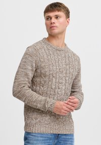 Lys brun kabelstrikket genser med rund hals og ribbestrikkede mansjetter. Brukt med blå denimjeans som viser et teksturert mønster.