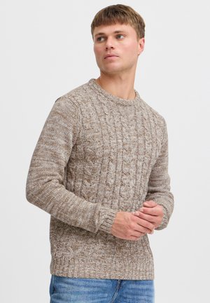 Hellbraunes, kabelstrickgestricktes Sweatshirt mit rundem Kragen und gerippten Bündchen. Getragen mit blauen Jeans aus Denim, die ein strukturiertes Muster zeigen.
