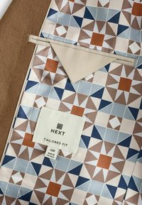 Brauner Stoff mit einem geometrischen Muster in Blau, Orange und Creme. Enthält ein Etikett für "NEXT TAILORED FIT" mit Größenangaben.