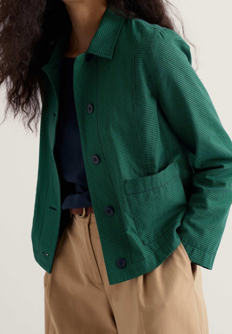 Femme portant une veste boutonnée à carreaux verts par-dessus un haut noir avec un pantalon taille haute beige et une ceinture marron, les mains dans les poches.