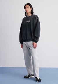 Giovane con capelli lunghi fino alle spalle, che indossa una felpa nera oversize, pantaloni larghi grigio chiaro e sneakers nere su un pavimento blu.