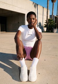 Jeune personne assise sur le sol en béton à l'extérieur près d'un bâtiment moderne, portant un t-shirt blanc, un short rose, des chaussettes blanches et des baskets.
