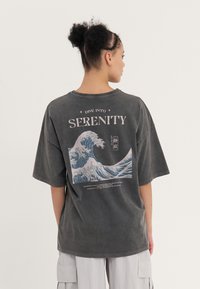 Graues, übergroßes T-Shirt mit einem Wellenmotiv und dem Wort "SERENITY" in Weiß auf dem Rücken bedruckt. Kurze Ärmel und runder Ausschnitt.