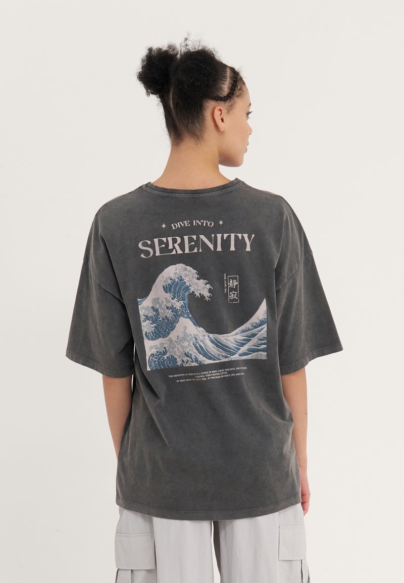 Graues, übergroßes T-Shirt mit einem Wellenmotiv und dem Wort "SERENITY" in Weiß auf dem Rücken bedruckt. Kurze Ärmel und runder Ausschnitt.