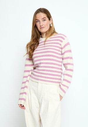 Mujer joven con cabello largo, vistiendo un suéter a rayas rosa y blanco y pantalones crema, de pie con una mano en el bolsillo.