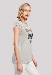 Grijze t-shirt met korte mouwen met een doodskopafbeelding en de tekst "Guns N Roses." Zacht materiaal met een comfortabele pasvorm. Gedragen met witte broek.