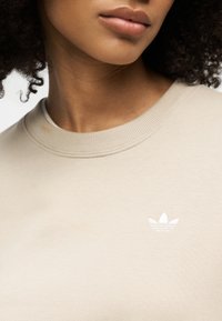 Femme aux cheveux bouclés portant un sweat-shirt beige col rond avec un petit logo blanc Adidas sur la poitrine.