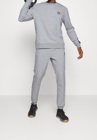 Grijze sweatshirt en bijpassende joggers gemaakt van zachte stof, met logopatch op de borst en zijstrepen. Zwarte sportieve schoenen met gumzolen.