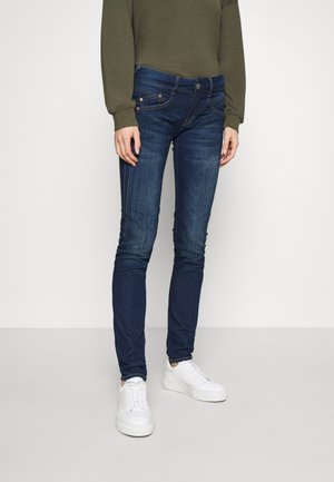 Osoba ubrana w ciemnoniebieskie dopasowane dżinsy, oliwkowozielony top z długim rękawem oraz białe sneakersy na platformie, stojąca na jednolitym tle.