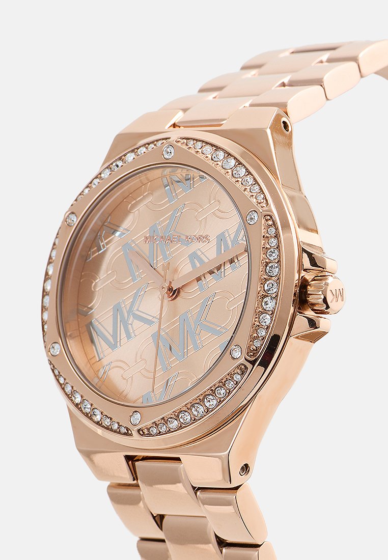 Montre Michael Kors en or rose avec un boîtier rond, accents en cristal, cadran à motifs présentant des logos MK entrelacés, et bracelet en maillons métalliques.