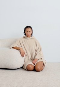 Beige hetteponcho laget av myk, teksturert stoff, med en frontlomme og avslappet passform, båret av en sittende figur på et lyst teppe.