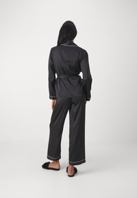 Svart satin pyjamas-set som består av en långärmad topp med bälte, kontrasterande piping och vida byxor. Modellen bär fluffiga svarta tofflor.