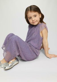 Fioletowy kombinezon z fałdami, wykonany z fakturowanej tkaniny, z rękawkami typu cap i błyszczącymi akcentami, w zestawie z lśniącymi srebrnymi baletkami. Pozycja siedząca.