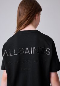 Hosszú barna hajú személy fekete pólóban, amelynek hátán az "ALLSAINTS", japán karakterek és a "LONDON" felirat látható.