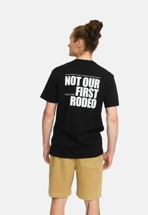 Zwarte katoenen t-shirt met op de achterkant vet witte tekst: "DIT IS NIET ONZE EERSTE RODEO" met daarboven kleine tekst. Korte mouwen.