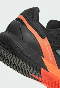 Μαύρο αθλητικό παπούτσι με πορτοκαλί διχτυωτό μέρος, υφή στο επάνω μέρος και καουτσούκ σόλα με σχέδιο. Περιέχει το κείμενο "DEFIANT SPEED" στο πλάι.