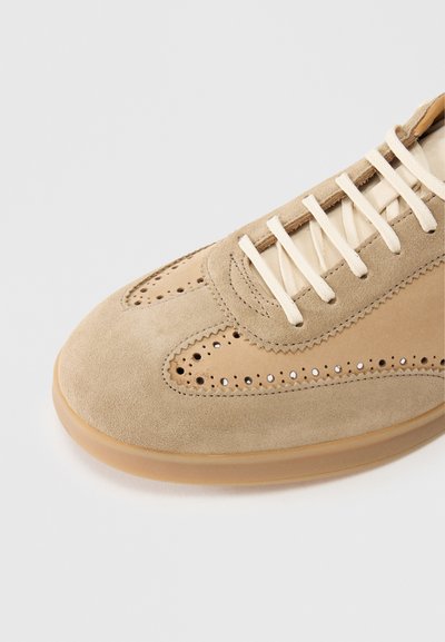 Baskets en daim beige avec accents perforés, lacets plats et semelle en caoutchouc. Texture lisse avec des détails de contraste de couleur claire.