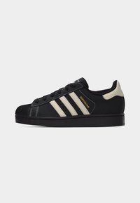 Μαύρο παπούτσι Adidas Superstar με μπεζ ρίγες, μαύρο καουτσούκ στη μύτη και το κείμενο «SUPERSTAR» σε χρυσό στο πλάι, εμφανιζόμενο σε προφίλ.