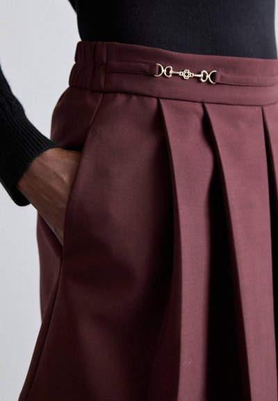 Jupe plissée bordeaux avec une taille élastique et des accents décoratifs dorés. Texture lisse avec des poches latérales. Vue rapprochée.