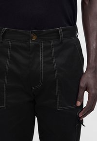 Pantalon noir avec des coutures contrastantes blanches et une fermeture par bouton, porté avec une chemise noire, montrant une partie de la main d'une personne sur le côté.