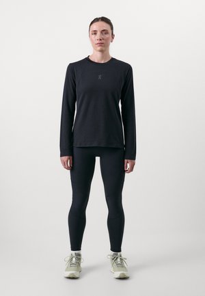 Haut noir à manches longues avec un col rond et un logo, associé à des leggings noirs ajustés et des chaussures de sport vert clair avec lacets.