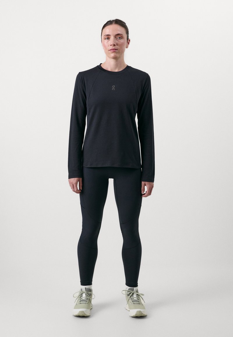 Schwarzes Langarmshirt mit Rundhalsausschnitt und Logo, kombiniert mit schmalen schwarzen Leggings und hellgrünen Sportschuhen mit Schnürsenkeln.