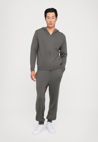 Grå treningssett bestående av en hettejakke med glidelås og ribbet mansjetter, samt matchende joggebukser. Stoffet har en glatt tekstur; kombinert med hvite joggesko.