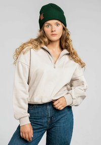 Beżowy sweter zapinany na zamek błyskawiczny z prążkowanym kołnierzem, w zestawie z ciemnoniebieskimi dżinsami. Dodatki to zielona dziana czapka z skórzaną metką.
