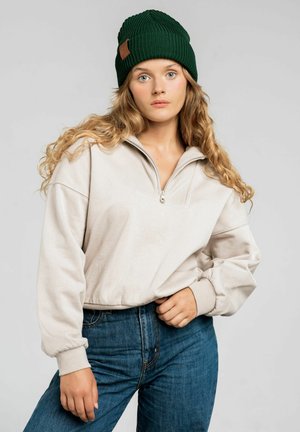 Beżowy sweter zapinany na zamek błyskawiczny z prążkowanym kołnierzem, w zestawie z ciemnoniebieskimi dżinsami. Dodatki to zielona dziana czapka z skórzaną metką.