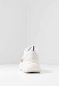 White sneaker with textured fabric upper, rubber sole, and subtle gold accents. Features a padded collar and pull tab for easy wear.